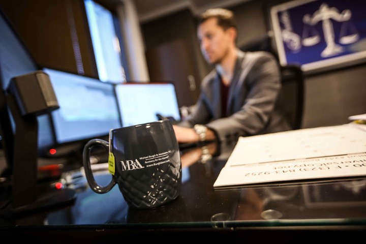 Close-up of Michael Burgis & Associates branded coffee mug on desk with blurred attorney working on catastrophic injury cases in background